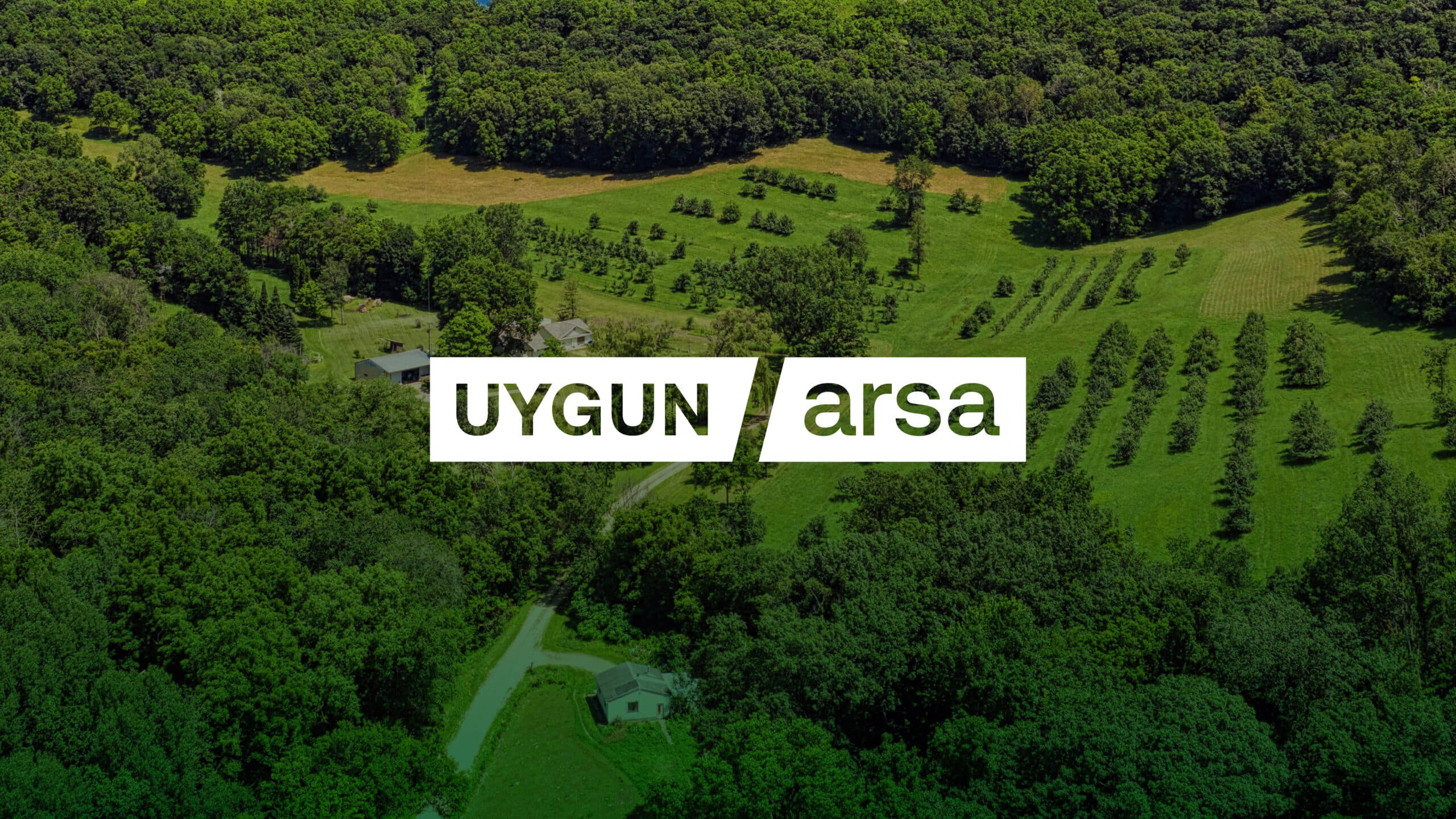 Aerial view of a lush, green landscape with dense forest, open fields with scattered trees, a winding dirt road, and a few small houses, overlaid with the text "UYGUN arsa".