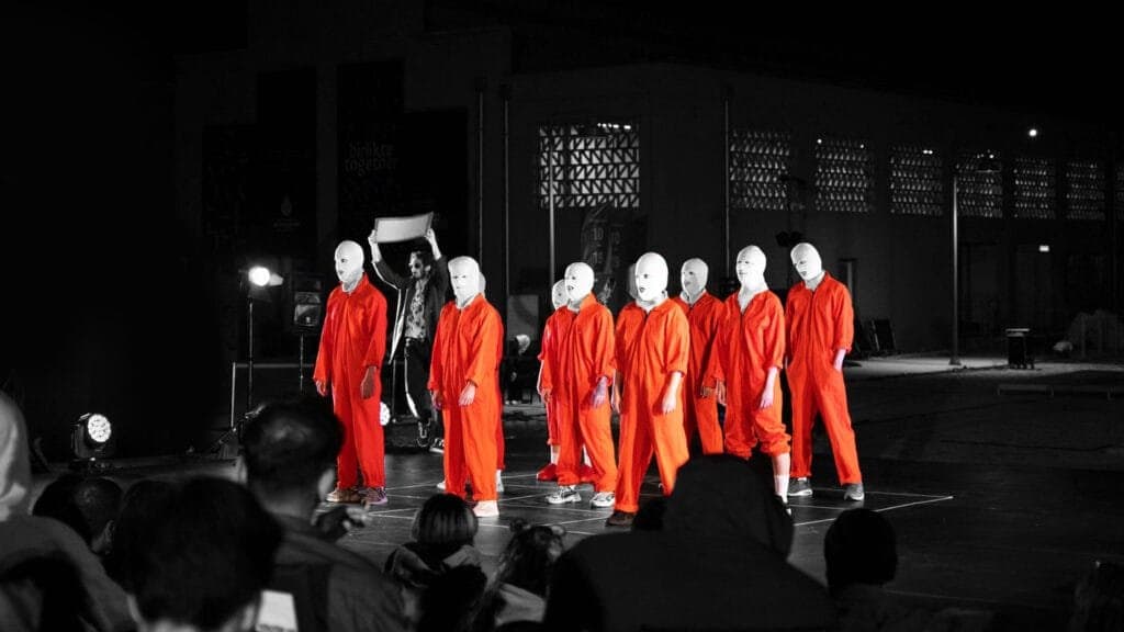 Performance art scene featuring a group of dancers in striking orange jumpsuits and white masks, standing in formation under dramatic stage lighting, with a mysterious figure in the background holding a sign. The audience watches intently, enhancing the immersive atmosphere of the event.